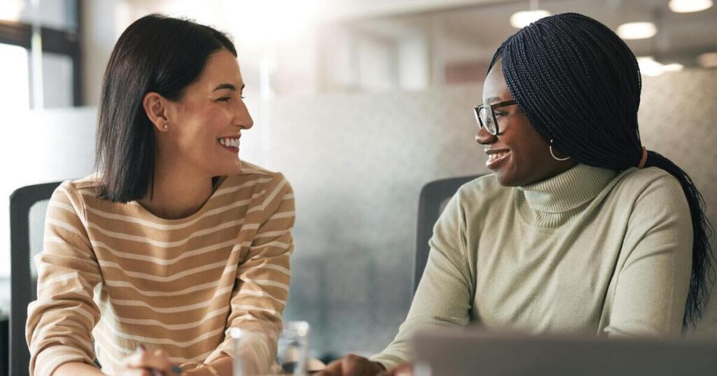 Two women sitting side by side at a worktable, talking and smiling at each other.