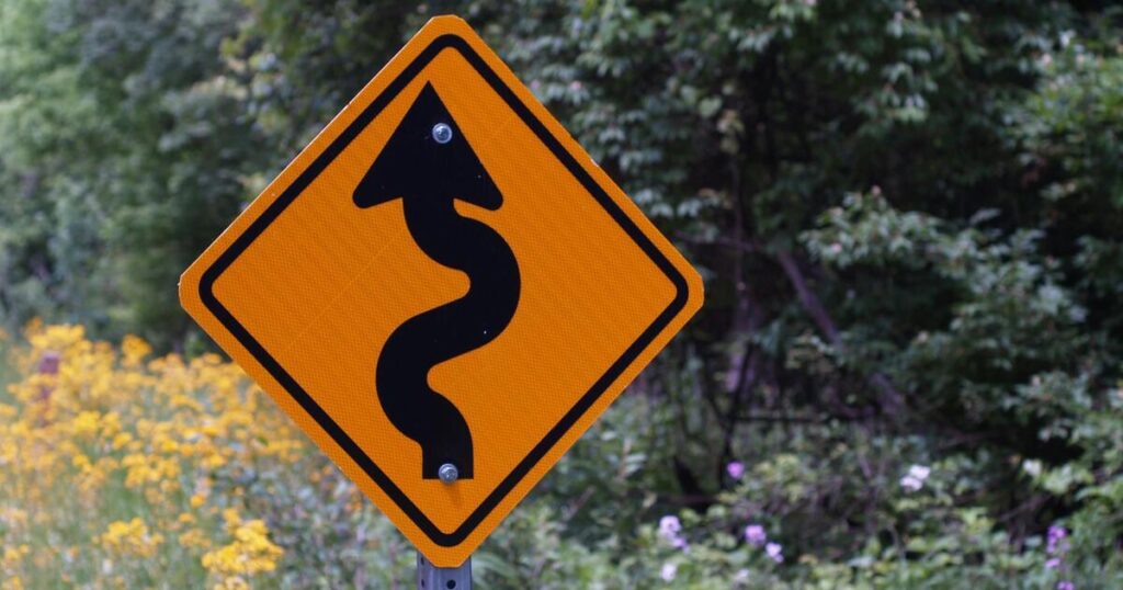 Curves-ahead roadsign: yellow sign with curving black arrow, forested background.