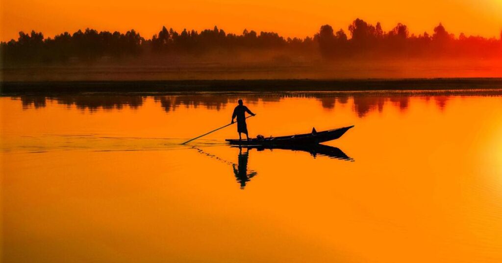 Silhouette of a person standing in a narrow boat, rowing across a calm lake at sunset, with orange sky and reflections on the water.