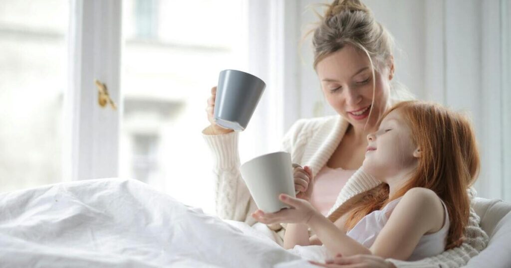 A mother and young daughter sit together on a bed near a bright window, smiling warmly as they hold mugs and enjoy a quiet morning moment together.