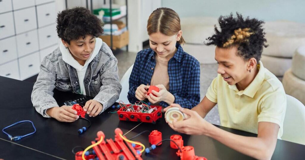 Three children sitting at a table working together on a hands-on robotics or STEM project with wires and small components, smiling and collaborating.